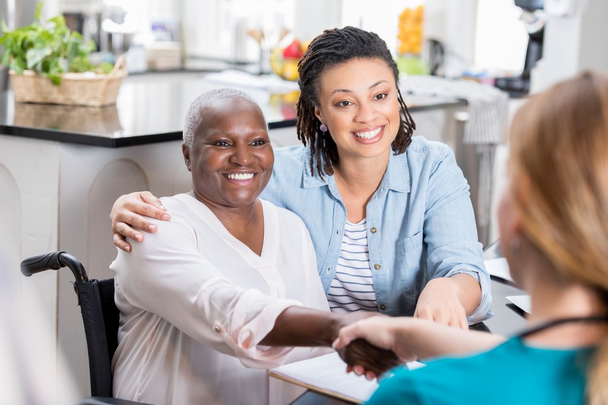 Caring mid adult woman talks with her mom's home healthcare nurse. The daughter has her arm around her mom. The senior woman is shaking the nurse's hand.